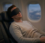 Man sleeping with a sleep mask on an airplane with a view of clouds outside the window.